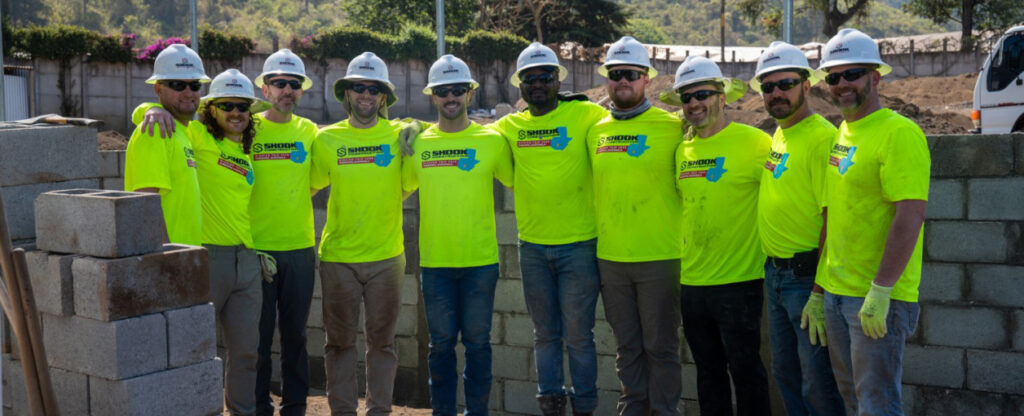 Ten people in bright yellow shirts and white hard hats stand arm-in-arm in front of a partially built concrete block wall at an outdoor construction site.