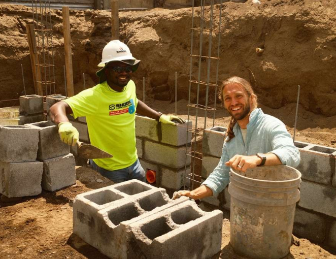 Two men smile while building a wall from cinder blocks at a construction site. One wears a hard hat, gloves, and a neon shirt; the other wears a light blue shirt and is holding a bucket. Dirt and rebar are visible in the background.