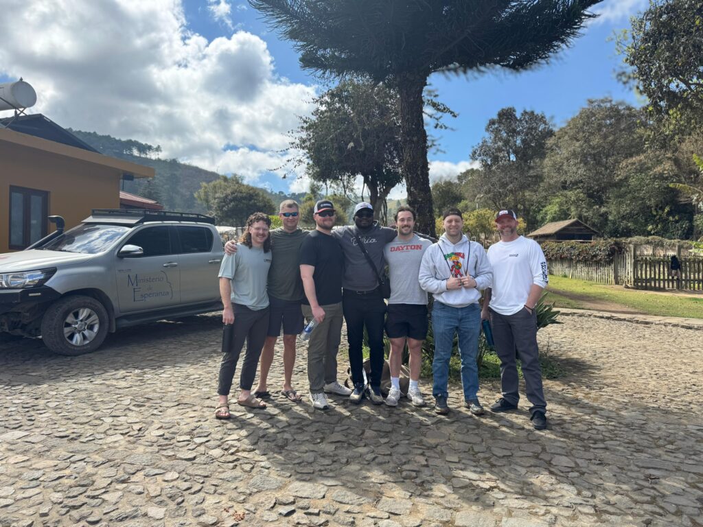 Seven people stand together on cobblestone, smiling in front of a tree and parked vehicle, with mountains, trees, and a house in the background under a partly cloudy sky.