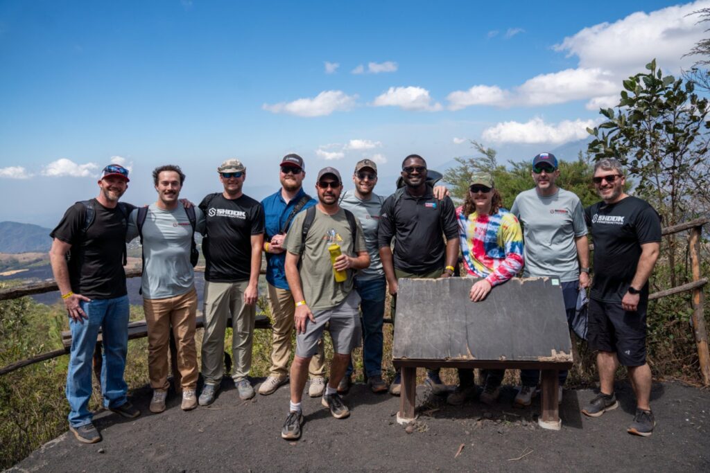 Ten people stand outdoors on a scenic overlook with blue sky and clouds. They are posing together, smiling, with trees and hills in the background and a wooden information sign in front of them.