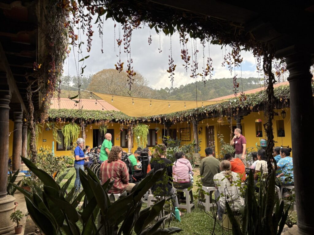 People gather in a lush, plant-filled courtyard with yellow walls, listening to speakers. String lights hang overhead, and greenery drapes from the roof. Mountains and trees are visible in the background under a cloudy sky.
