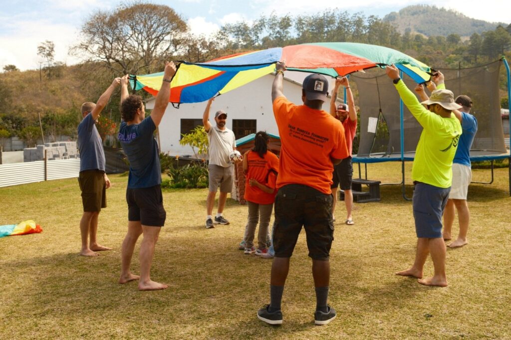 A group of people stand in a circle outdoors, lifting a colorful parachute. Children and adults participate, surrounded by grass, trees, and mountains under a partly cloudy sky.