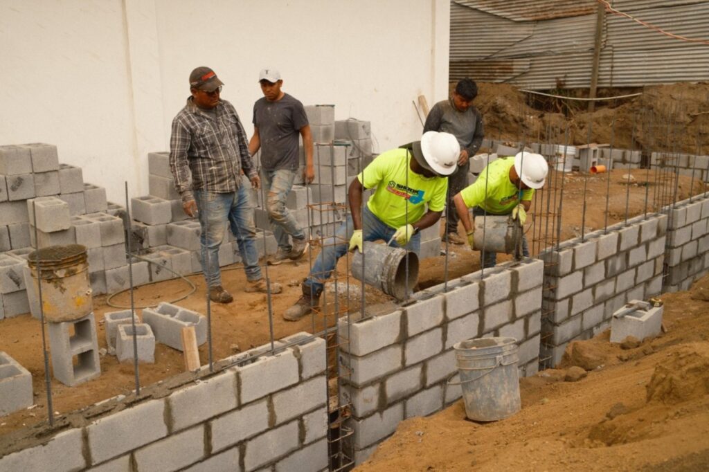 Two construction workers in yellow shirts pour cement into a row of concrete blocks, while three others observe nearby at a building site.