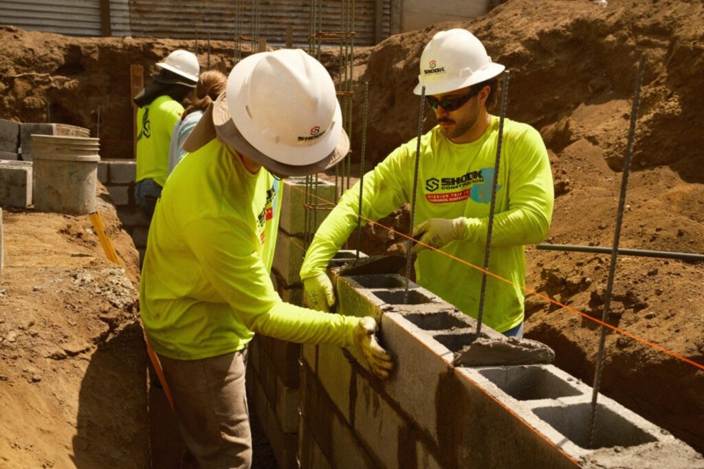 Two construction workers in yellow shirts and helmets build a concrete block wall at a construction site, measuring and aligning the blocks with an orange string line. Dirt and tools are visible in the background.