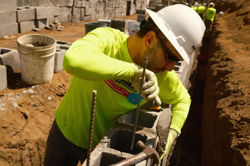 A construction worker in a white hard hat and neon shirt uses a tool to adjust rebar in concrete blocks, building a wall at a construction site with others working in the background.