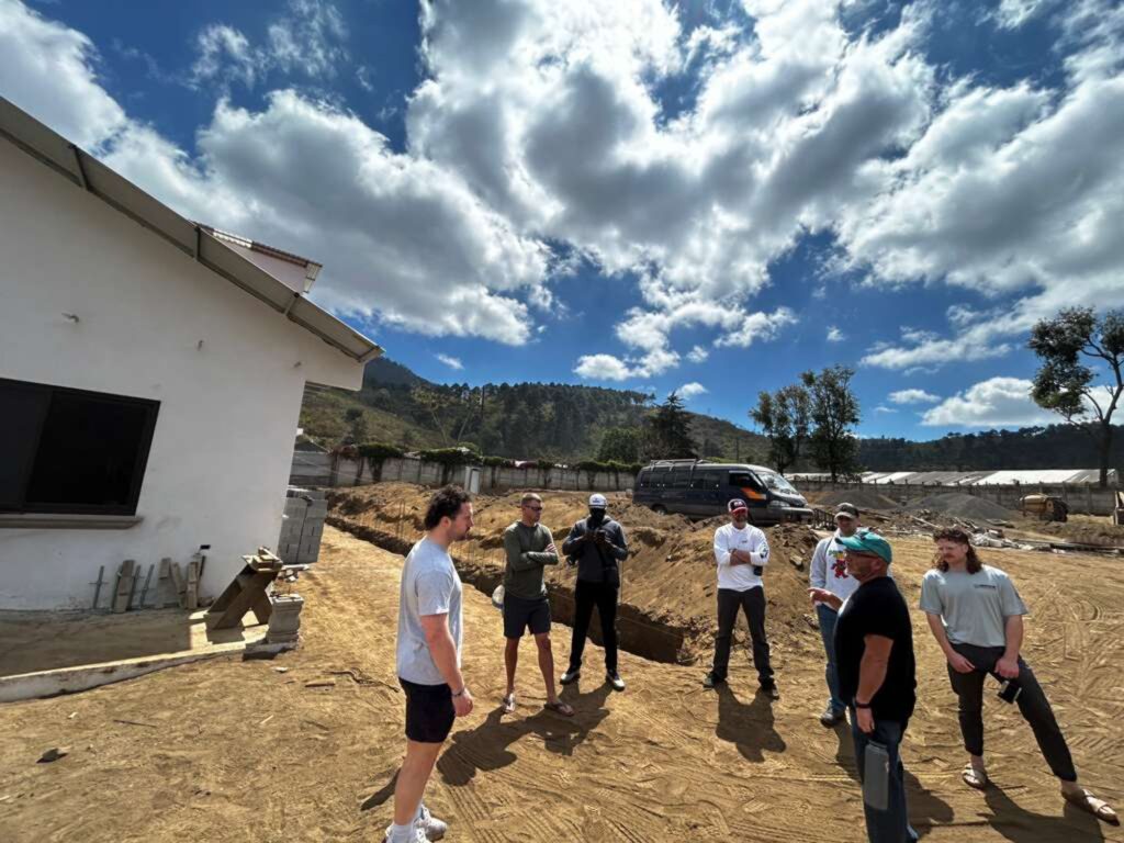 A group of people stand in a dirt area near a white building, talking under a partly cloudy sky with hills and trees in the background. A van is parked nearby and construction materials are visible on the ground.