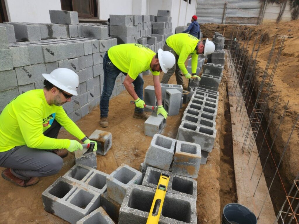 Three workers in neon yellow shirts and white hard hats lay cement blocks along a building foundation, with steel rods and sand visible at the construction site.
