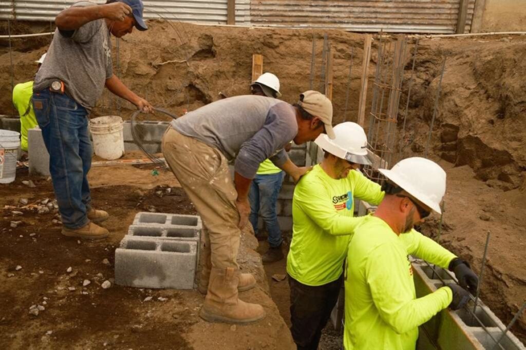 Five construction workers in safety gear lay concrete blocks and rebar at a building site, surrounded by dirt and building materials. Two workers are bent over, focusing closely on their tasks.