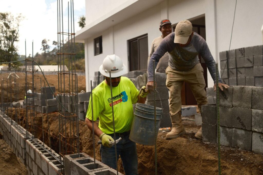 Two construction workers build a cinder block wall; one lifts a bucket while the other moves a block. Both wear helmets and safety gear, working next to a white building under construction.