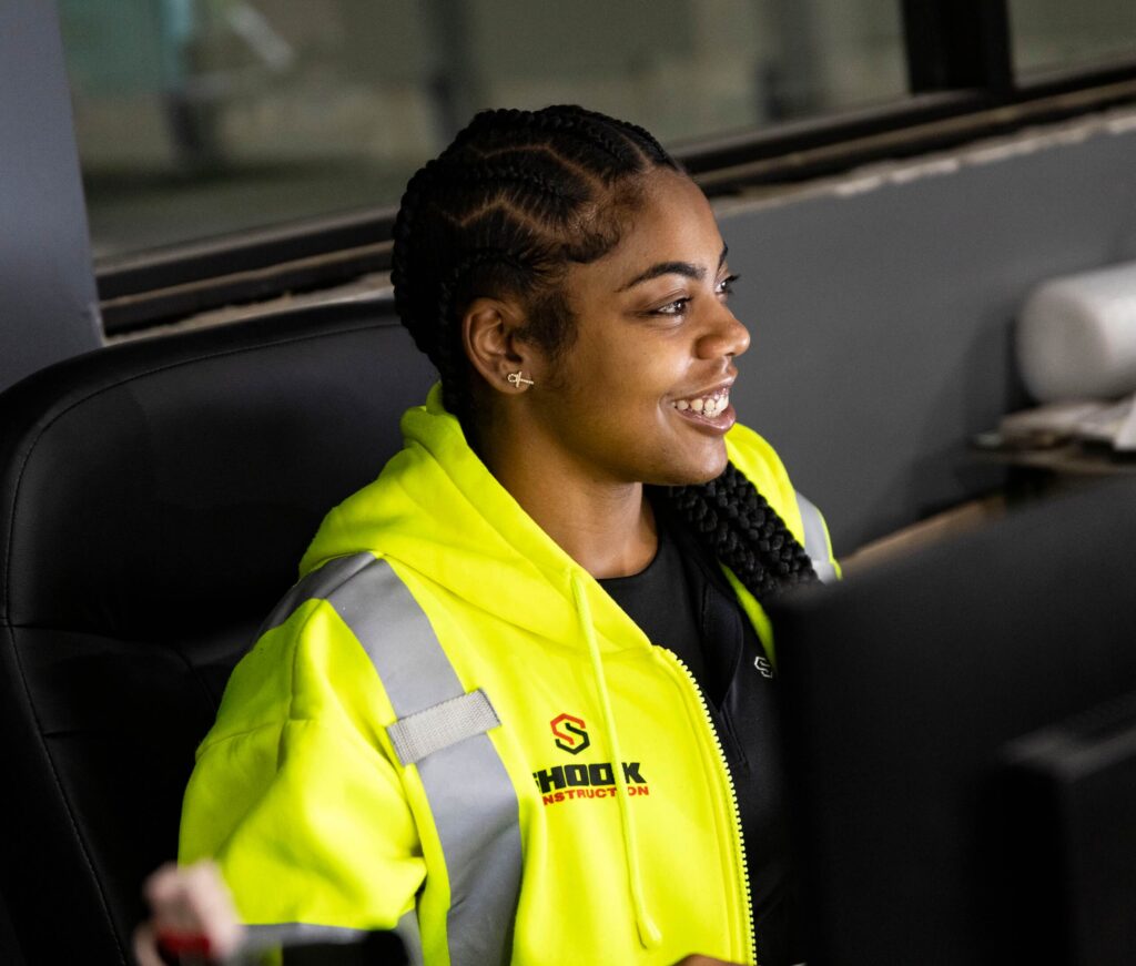 A woman wearing a neon yellow reflective jacket with SHOOK CONSTRUCTION sits at a desk, smiling and looking at a computer screen.