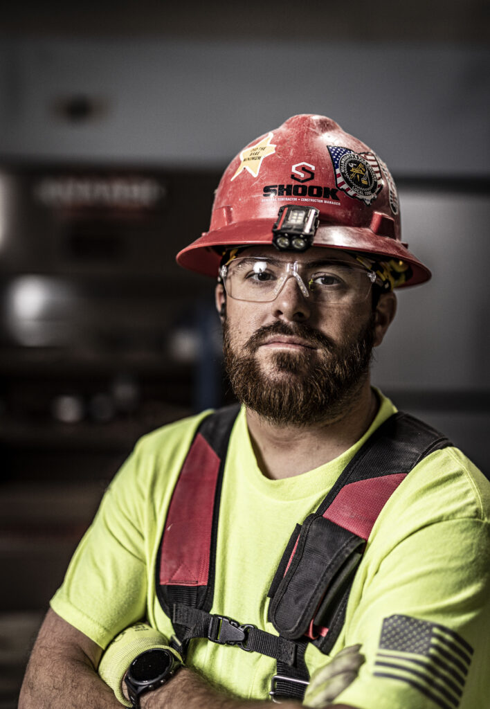 A construction worker wearing a red hard hat, safety glasses, neon yellow shirt, and harness stands with arms crossed, looking at the camera.