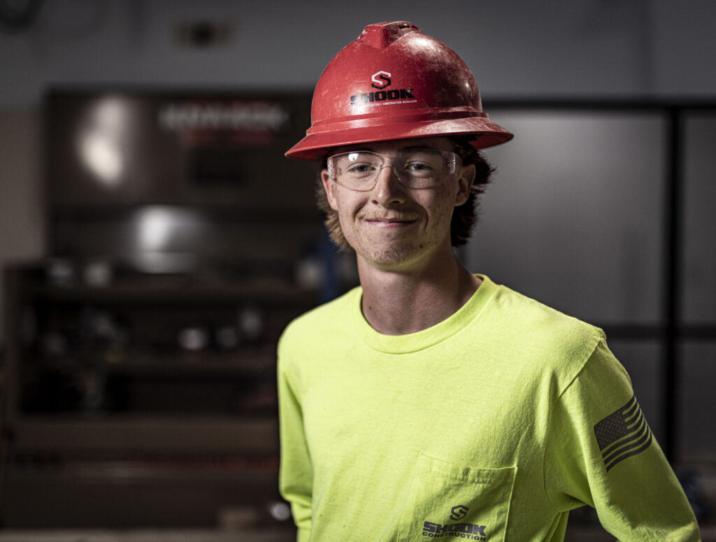 Young person wearing a red hard hat, safety glasses, and a neon yellow shirt with a logo, standing indoors in a workshop or industrial setting, smiling at the camera.