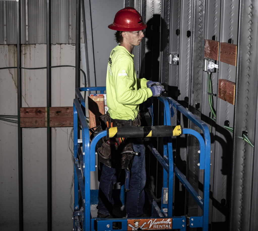 A construction worker in a red hard hat and yellow shirt stands on a blue lift platform, working on a metal wall indoors. He wears a tool belt and gloves, facing the wall with wiring and wooden boards.