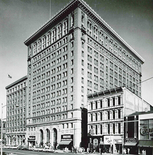 Black-and-white photo of a tall, historic, multi-story building on a city street corner, with shops and pedestrians at street level and classic architectural details above.