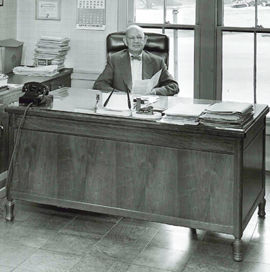 An elderly man in a suit sits at a large wooden desk with papers, a telephone, and office supplies, in an office with tall windows behind him.