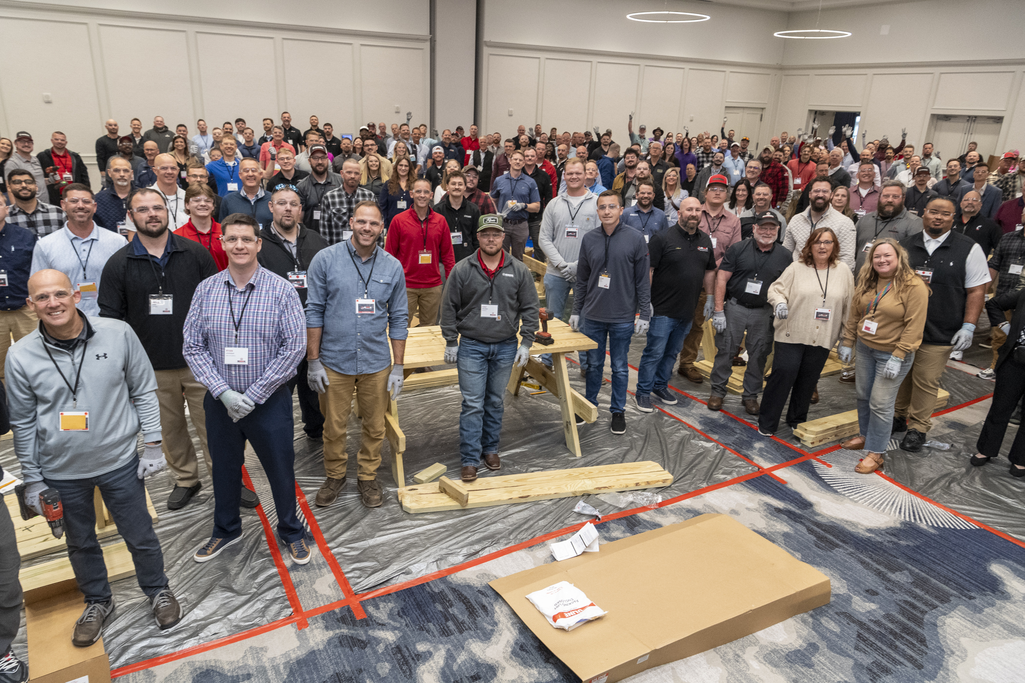 Large group of people wearing name badges stand and smile in a conference room with construction materials on the floor, facing the camera.