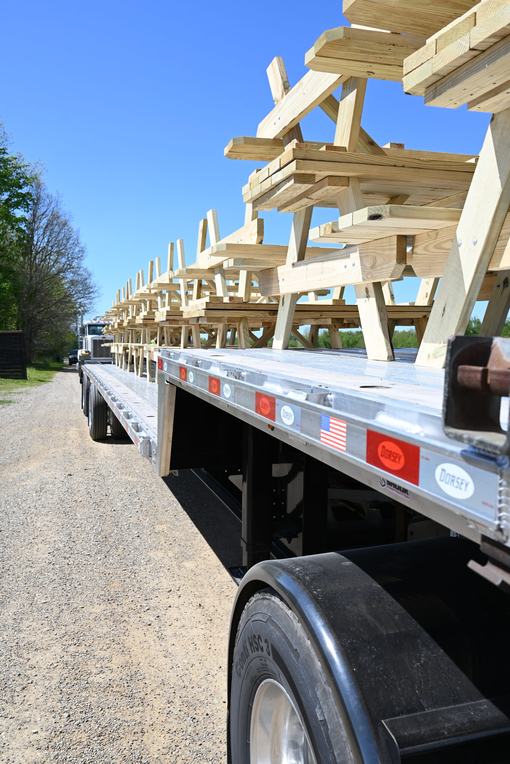 Flatbed trailer loaded with multiple unpainted wooden picnic tables, stacked and secured, parked on a gravel road under a clear blue sky.