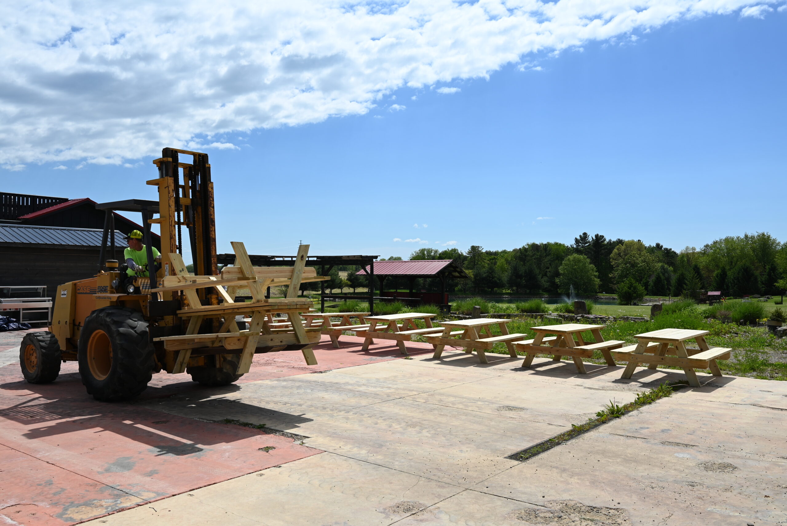 A forklift carries stacked wooden picnic tables across a concrete area outdoors, with more picnic tables lined up nearby and greenery in the background under a partly cloudy sky.