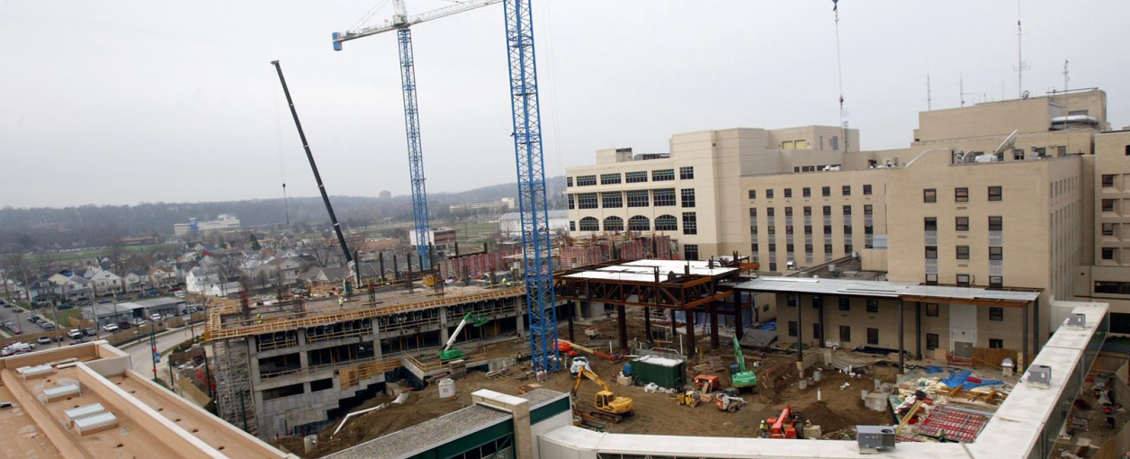 A large construction site with cranes and machinery, adjacent to a multi-story hospital building on a cloudy day. Workers and vehicles are visible, with steel framework and concrete structures in progress.