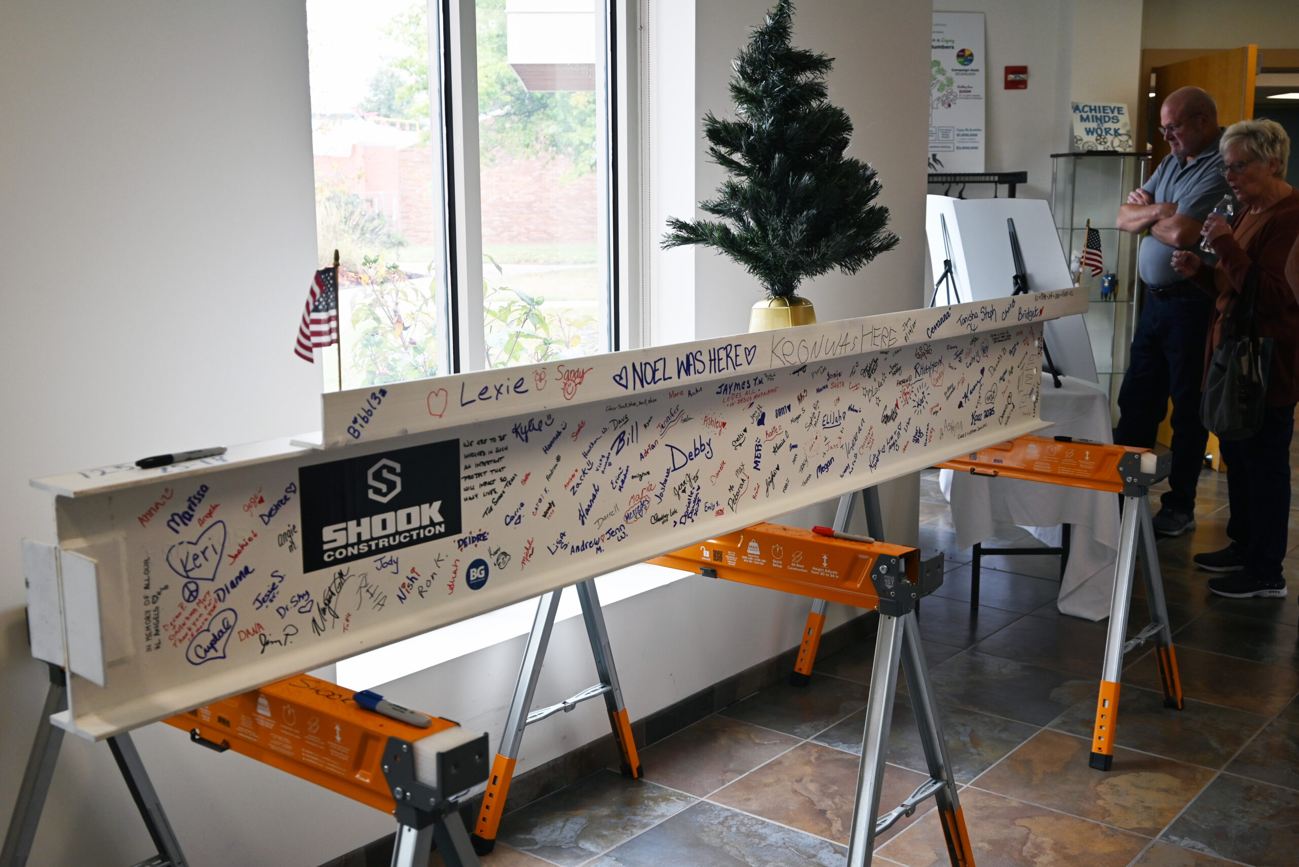 A construction beam signed with messages and names is displayed indoors on sawhorses near a window, small tree, and two people. A Shook Construction logo is visible on the beam.