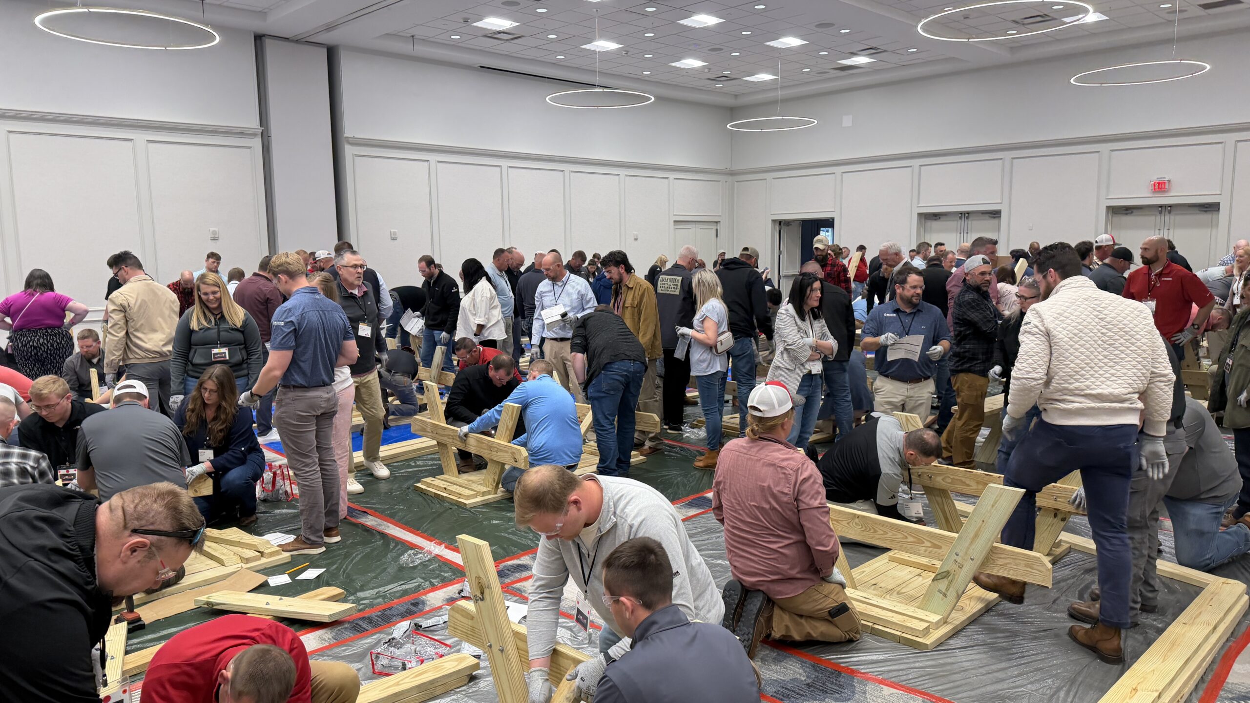 A large group of people inside a conference room assembling wooden structures on plastic-covered floors, with some standing and others kneeling, all appearing engaged in a team-building or workshop activity.