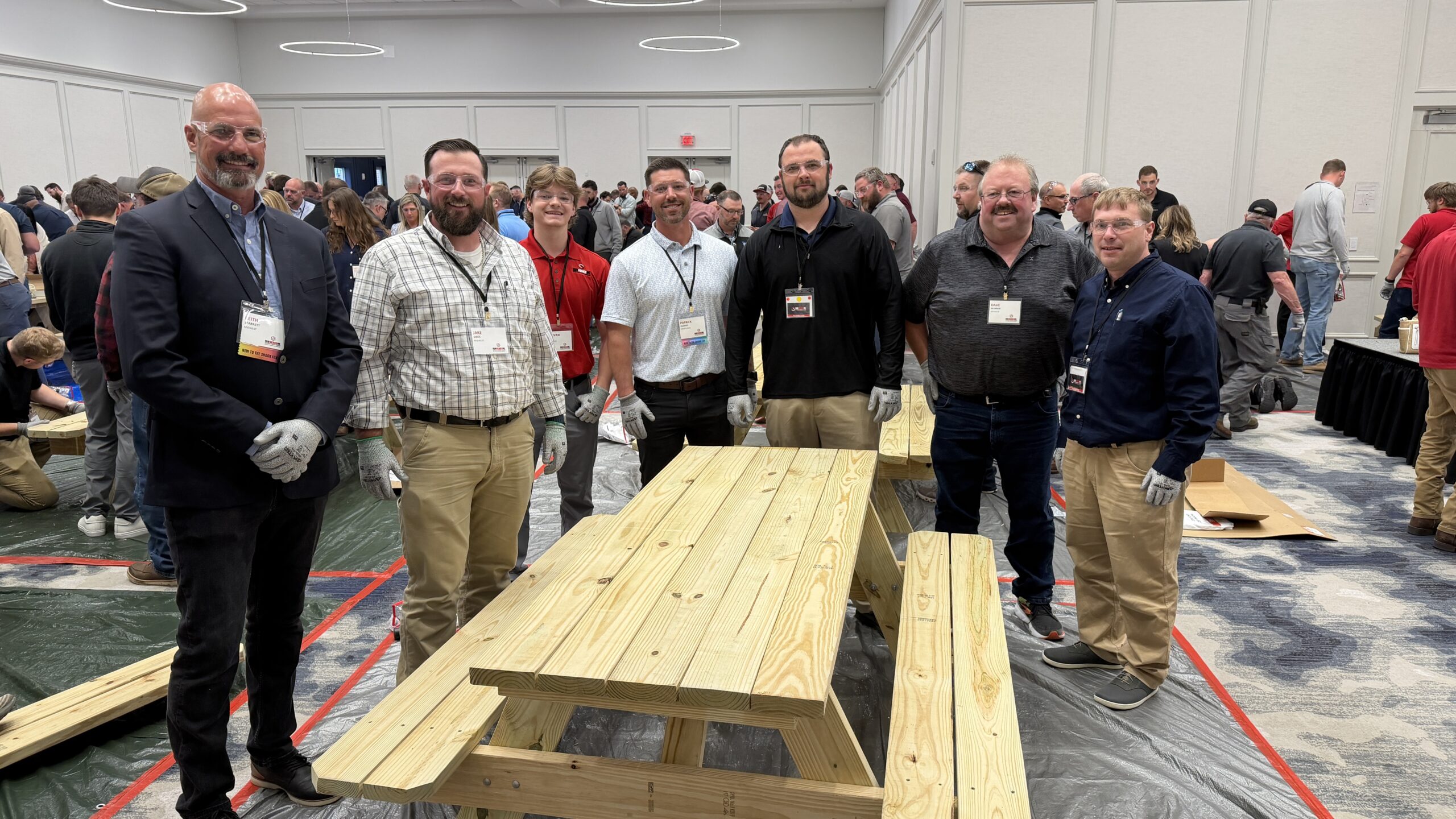 Seven men wearing gloves and name badges stand around a new wooden picnic table in a conference room, with others working in the background.