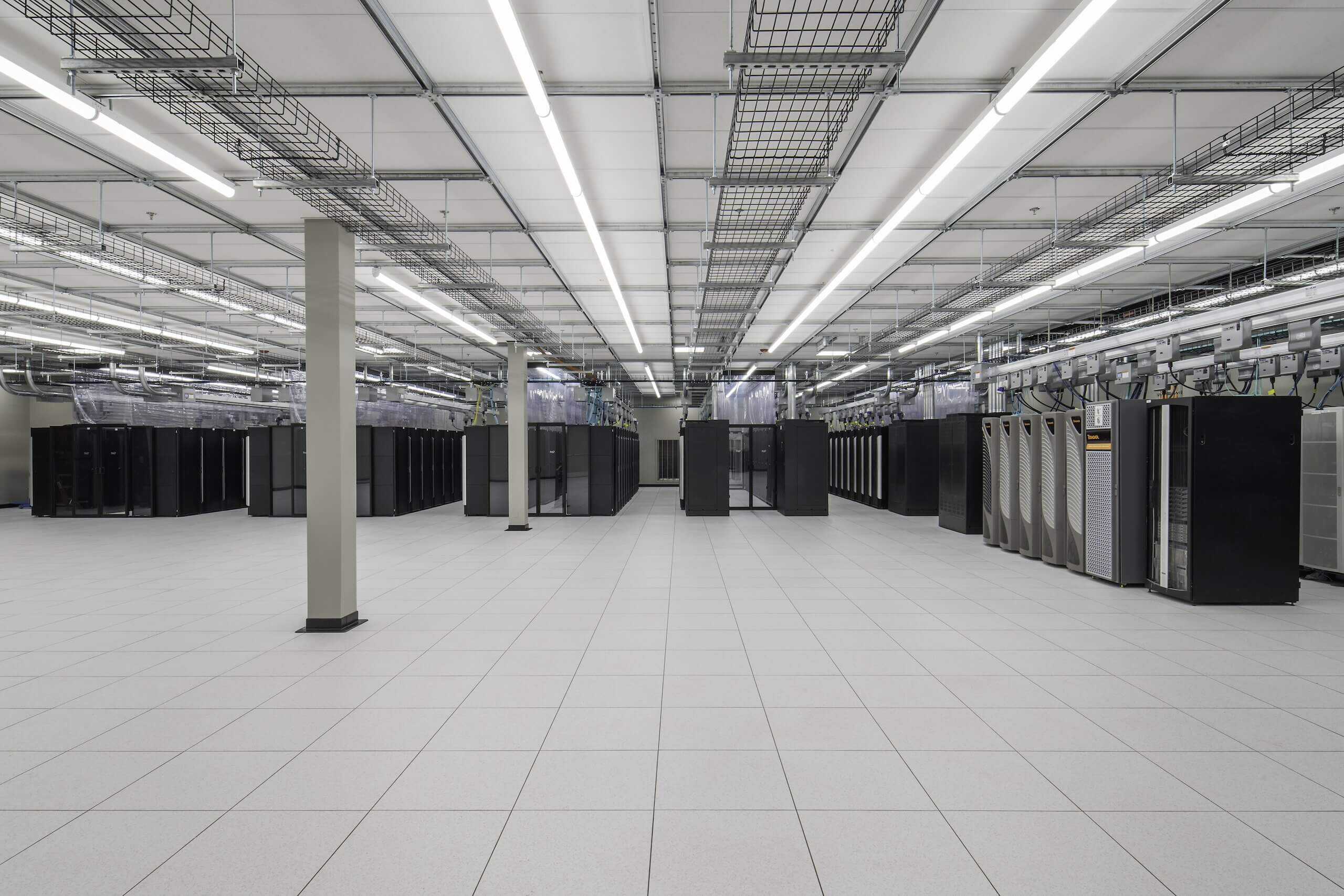 A large, modern data center room with rows of black server racks, overhead cable trays, and bright fluorescent lighting, all on a clean white tiled floor.