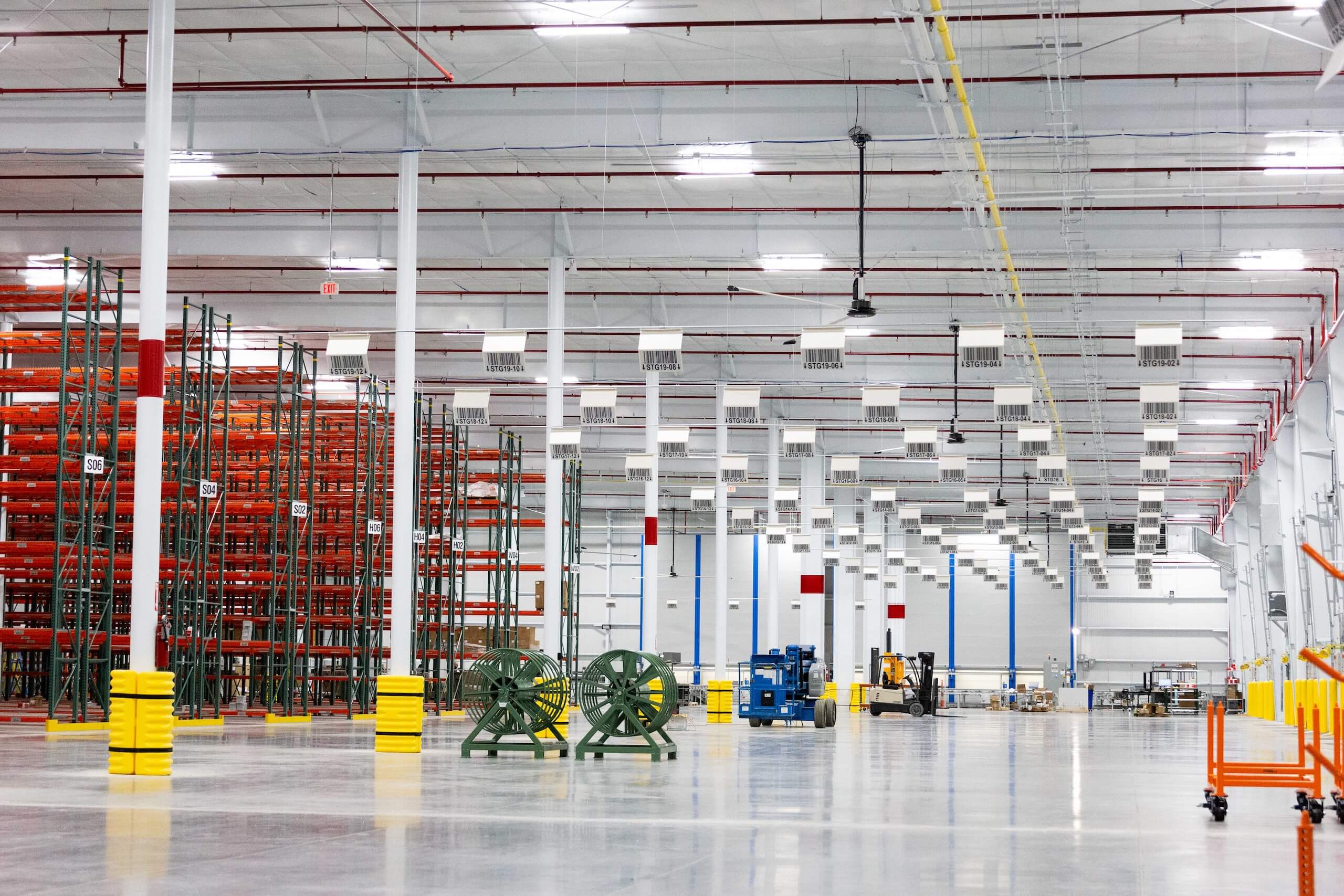 Large, brightly lit warehouse interior with empty red and green storage racks on the left, industrial equipment, and yellow safety barriers on the support columns.