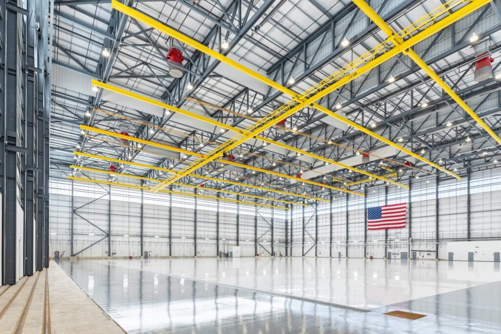 A large, empty aircraft hangar with polished floors, exposed steel beams, yellow safety rails, overhead lights, and a U.S. flag hanging on the back wall. The space is spacious and brightly lit.