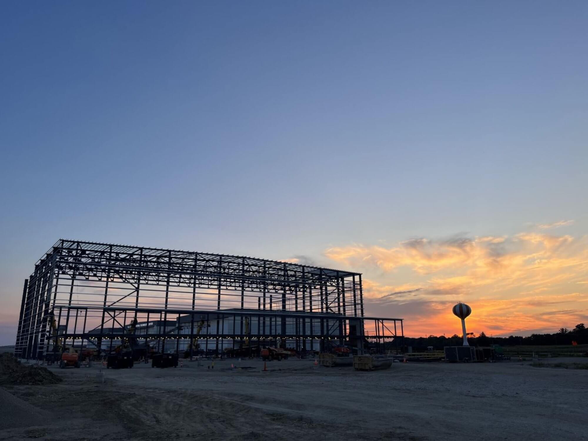 Steel frame of a large building under construction at sunset, with orange clouds in the sky and a water tower in the background.