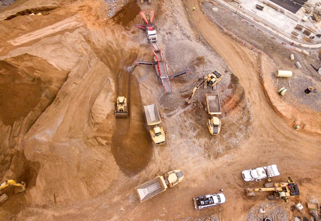 Aerial view of a construction site with dump trucks, excavators, and heavy machinery moving dirt and gravel across the area.