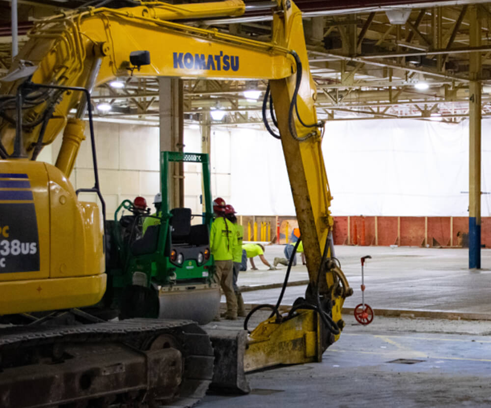 A yellow Komatsu excavator is parked indoors. Workers in safety gear are using tools and equipment to work on the concrete floor of a large industrial building.