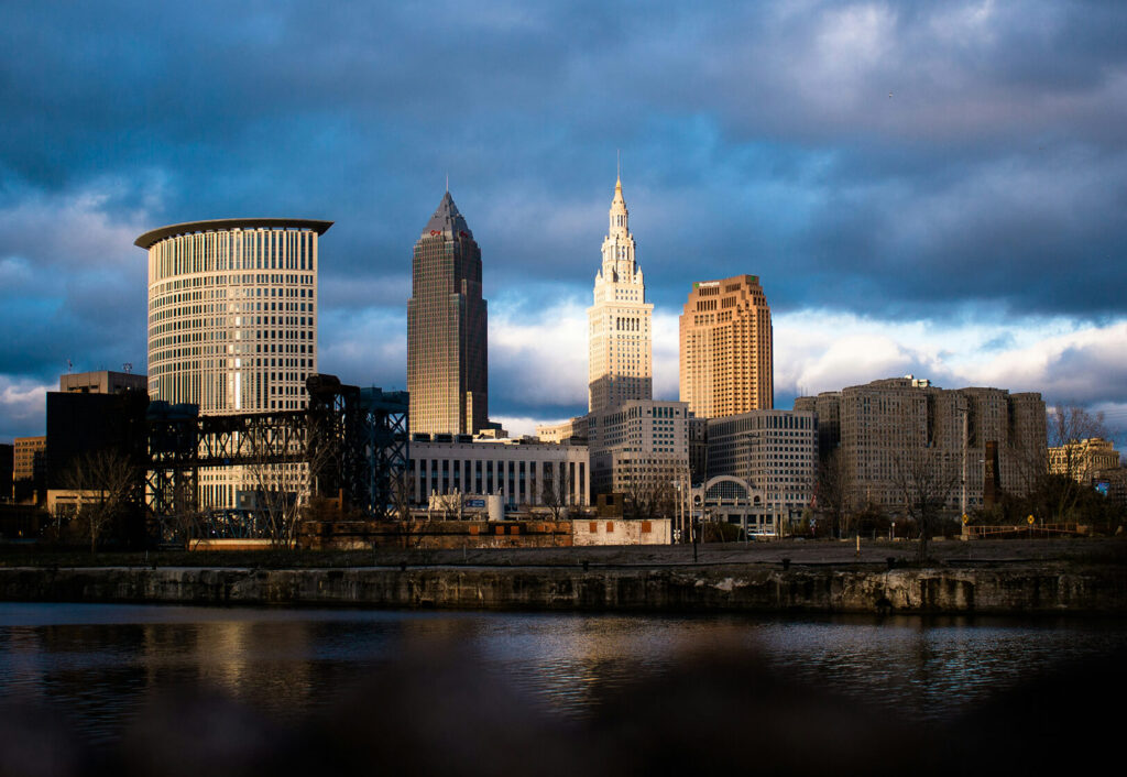 Cleveland skyline at sunset with tall buildings, including the Key Tower and Terminal Tower, under a dramatic, partly cloudy sky, reflected in the water below.