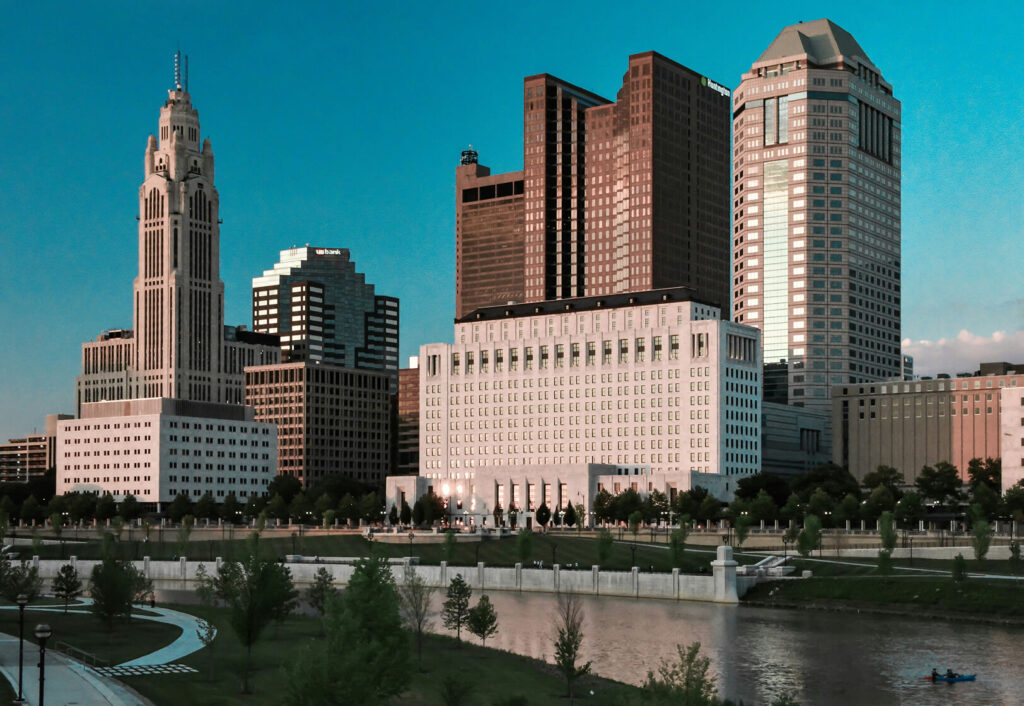A city skyline with tall buildings near a river, green trees along the water, and a blue sky in the background.