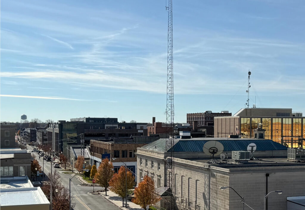 A cityscape shows a street lined with autumn trees, modern and historic buildings, a water tower in the distance, and a clear sky above with wispy clouds.
