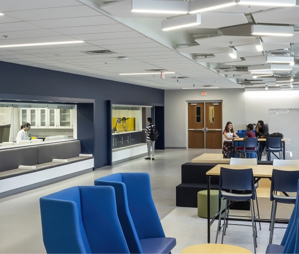 Modern common area with blue chairs, a group of people talking at a table, a student walking by lockers, and a person working behind a counter in a well-lit, spacious interior.