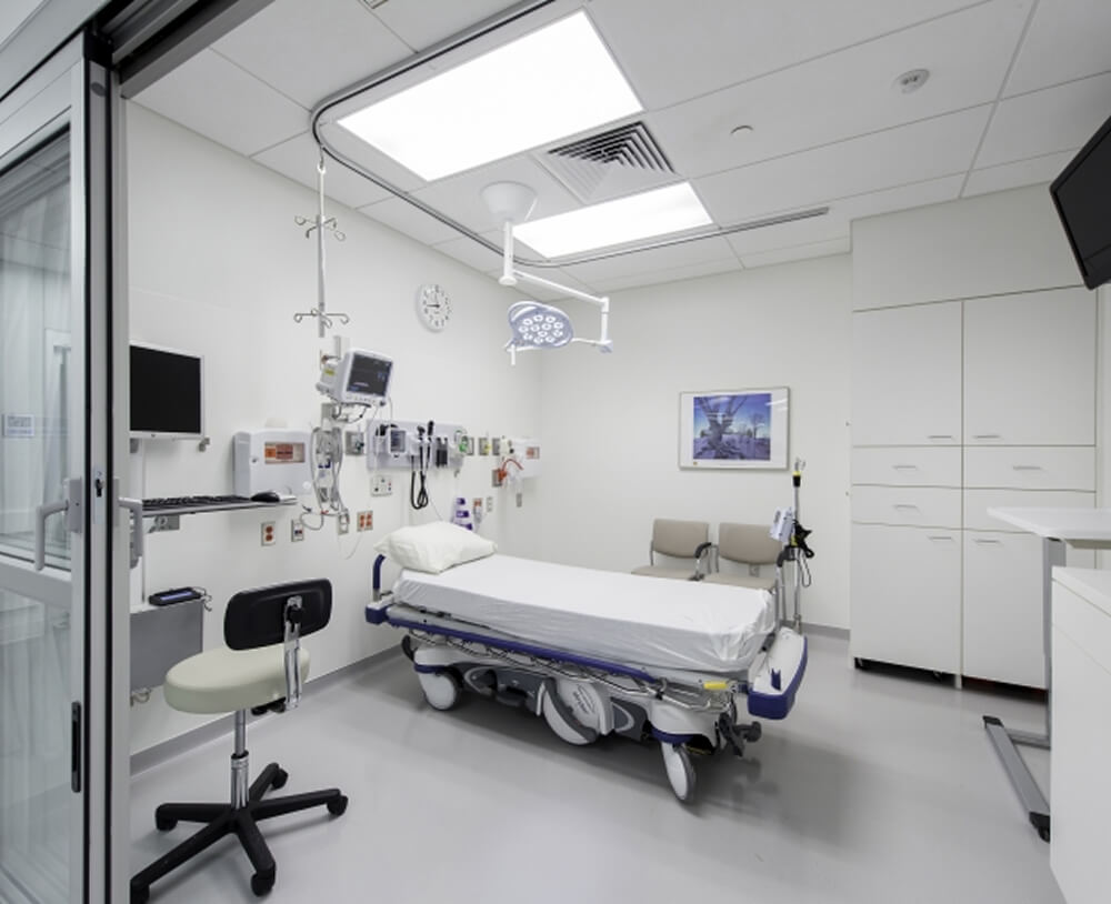 A modern, clean hospital room with a patient bed, medical equipment on the walls, a monitor, chairs, cabinets, and bright lighting. The room is empty and well-organized, with a photo on the wall and a large overhead light.