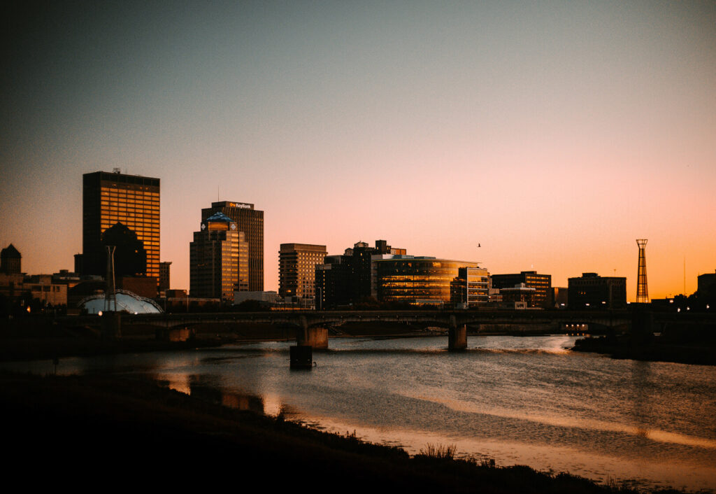 Downtown city skyline at sunset with modern buildings, a bridge over a calm river, and soft orange light reflecting on the water.