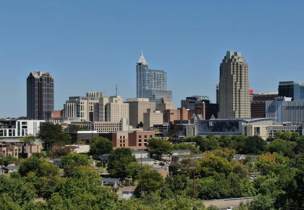 Downtown city skyline with modern and historic tall buildings under a clear blue sky, surrounded by green trees and smaller houses in the foreground.