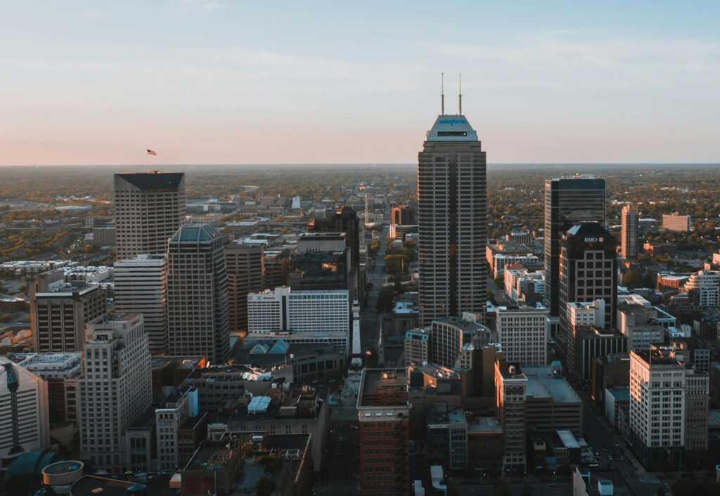 Aerial view of a city skyline with tall skyscrapers, office buildings, and streets below at sunset. The sky is clear with warm sunlight casting shadows over the urban landscape.