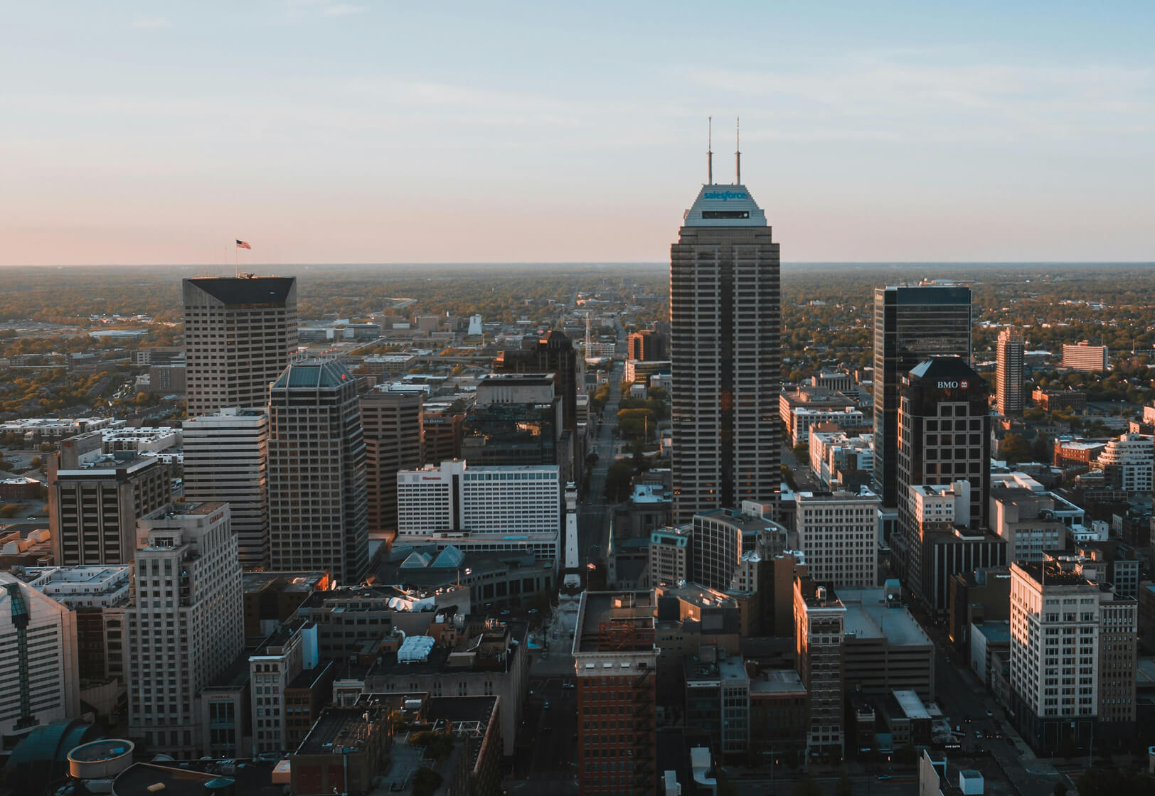 Aerial view of a city skyline with tall skyscrapers, office buildings, and streets below at sunset. The sky is clear with warm sunlight casting shadows over the urban landscape.