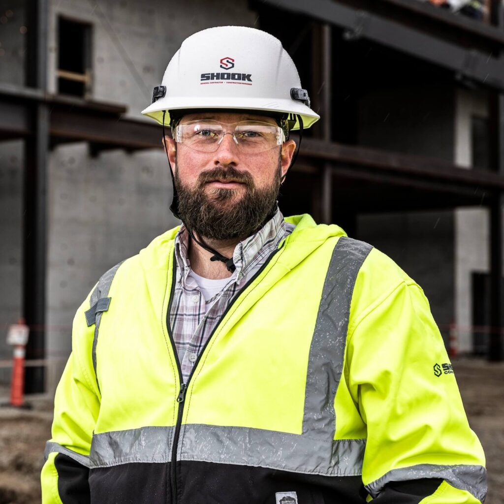 A bearded man wearing a white hard hat, safety glasses, and a neon yellow safety jacket stands at a construction site with steel beams and concrete in the background.