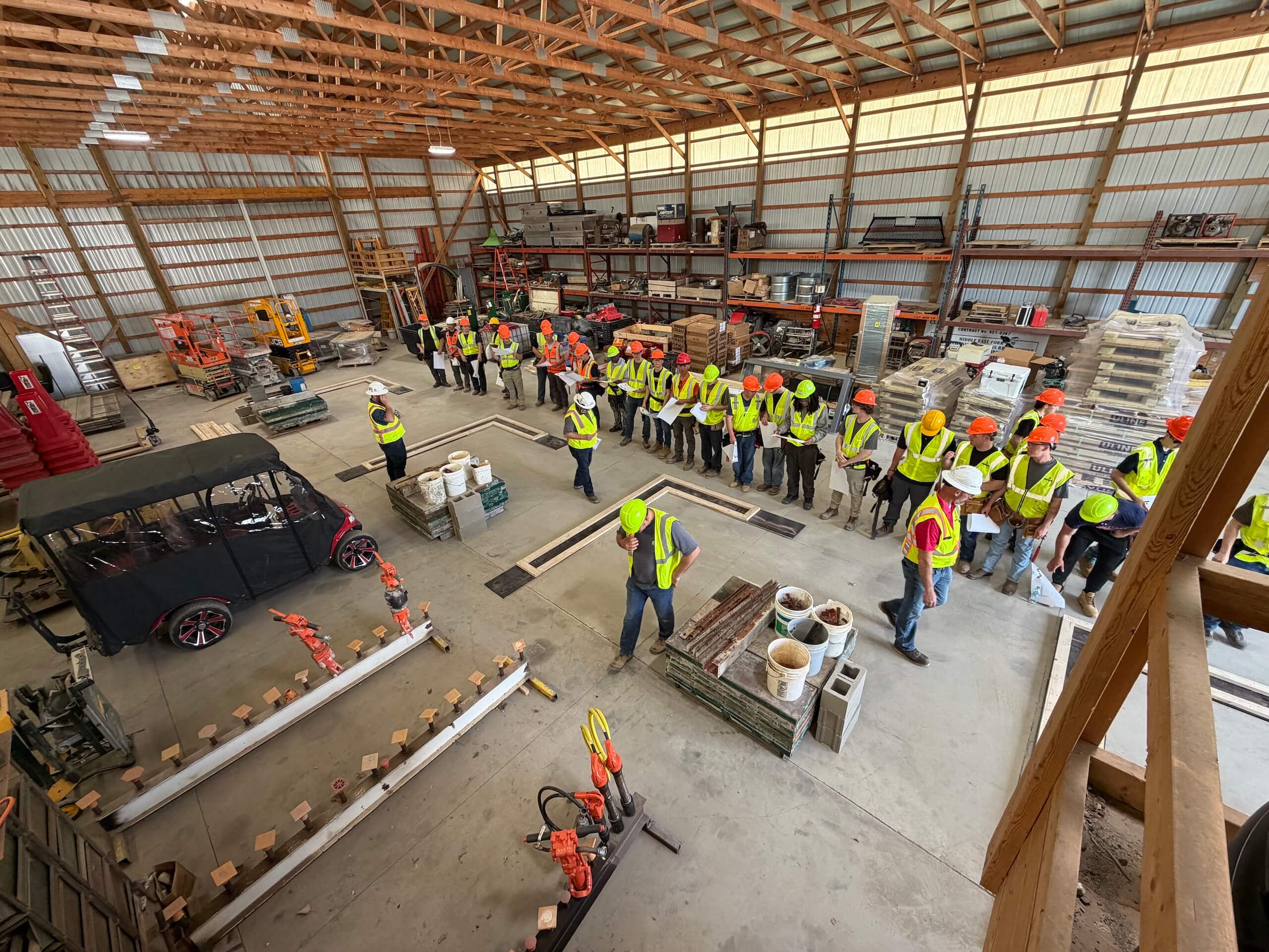 A group of people wearing safety vests and hard hats stand in a semicircle inside a large warehouse with construction materials and equipment.