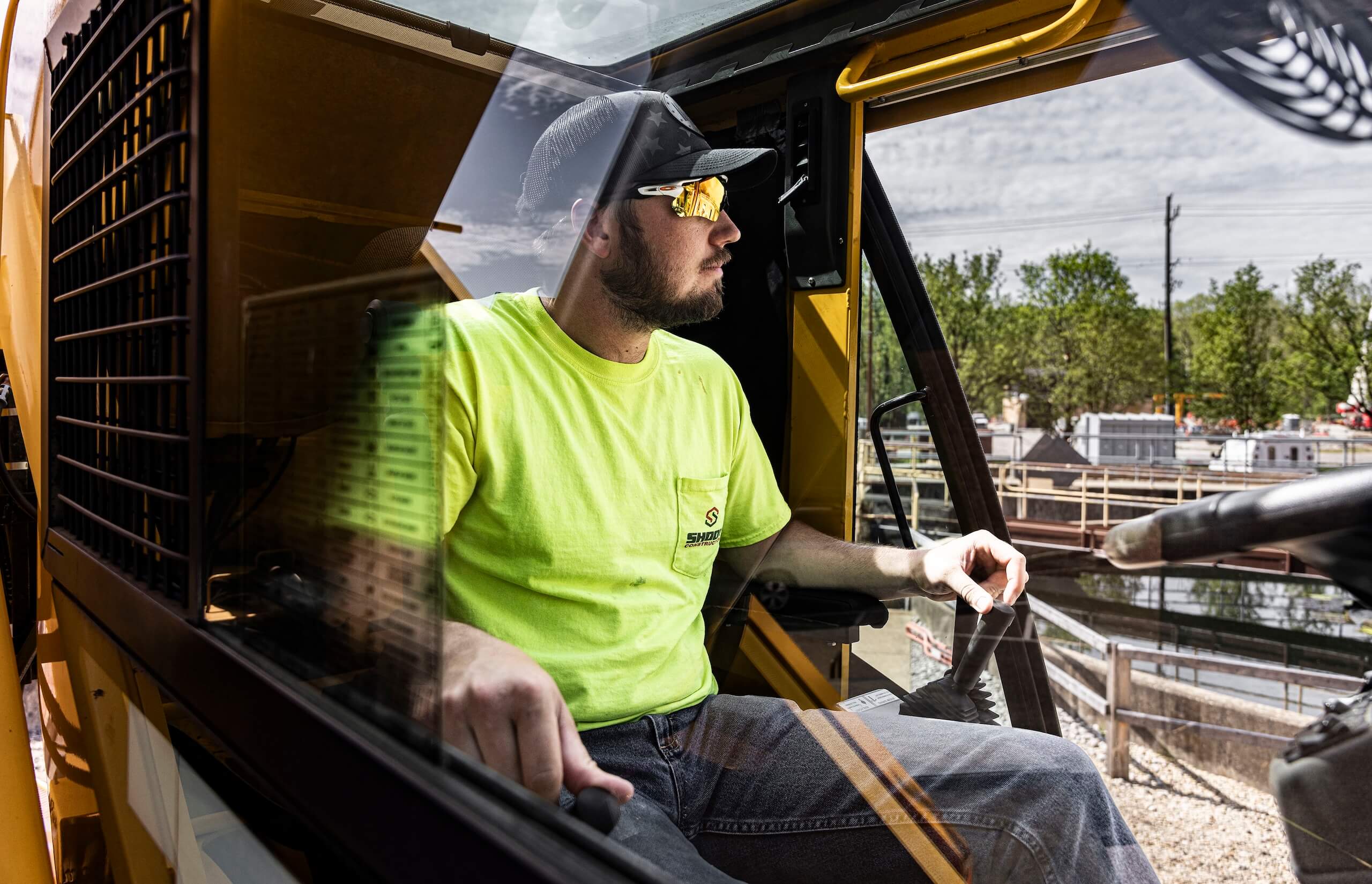 A man in a neon yellow shirt and sunglasses sits in the cab of construction equipment, looking out the window at a work site with trees and utility poles in the background.