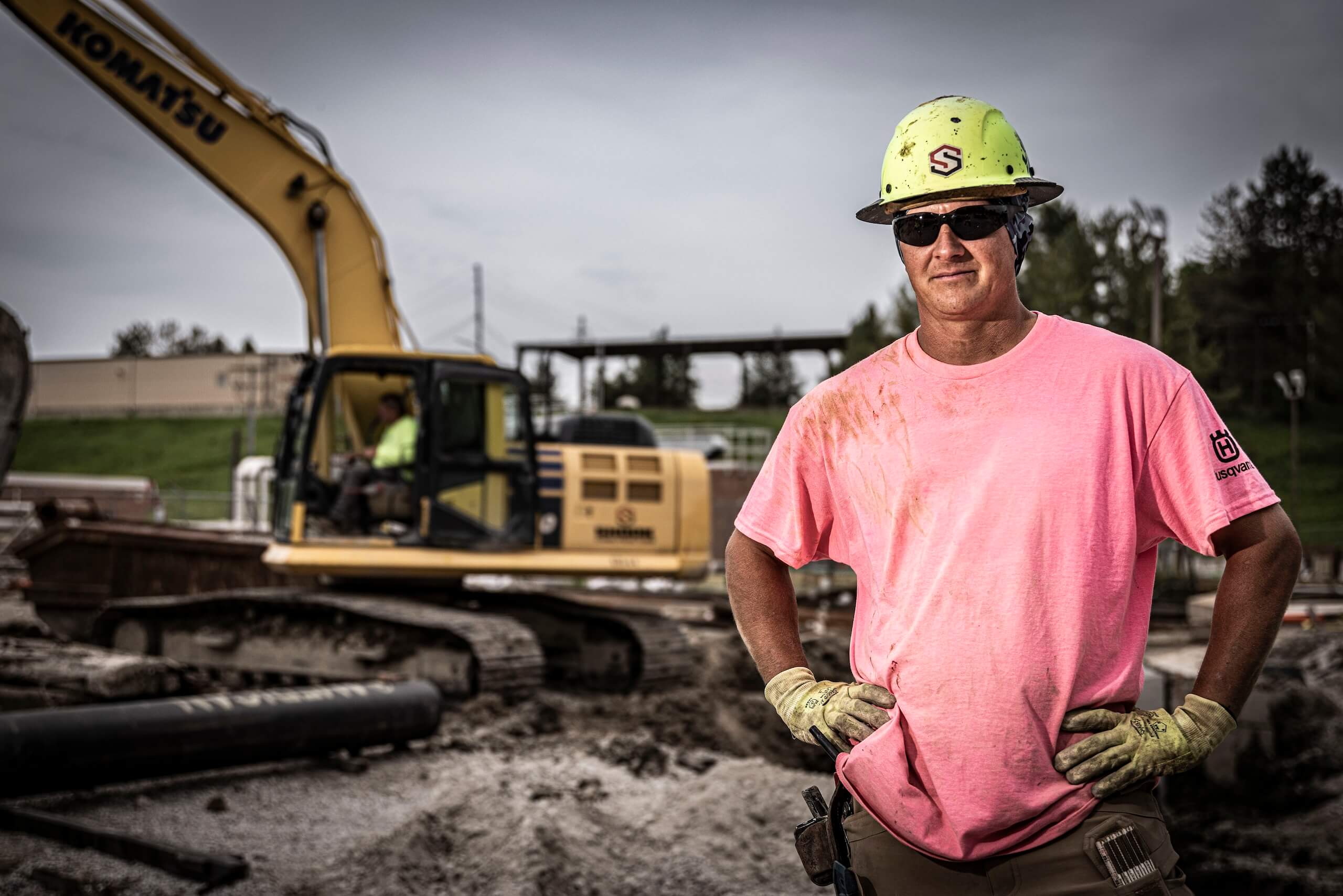 A construction worker in a pink shirt and yellow hard hat stands with hands on hips at a muddy job site, with an excavator and another worker in the background.