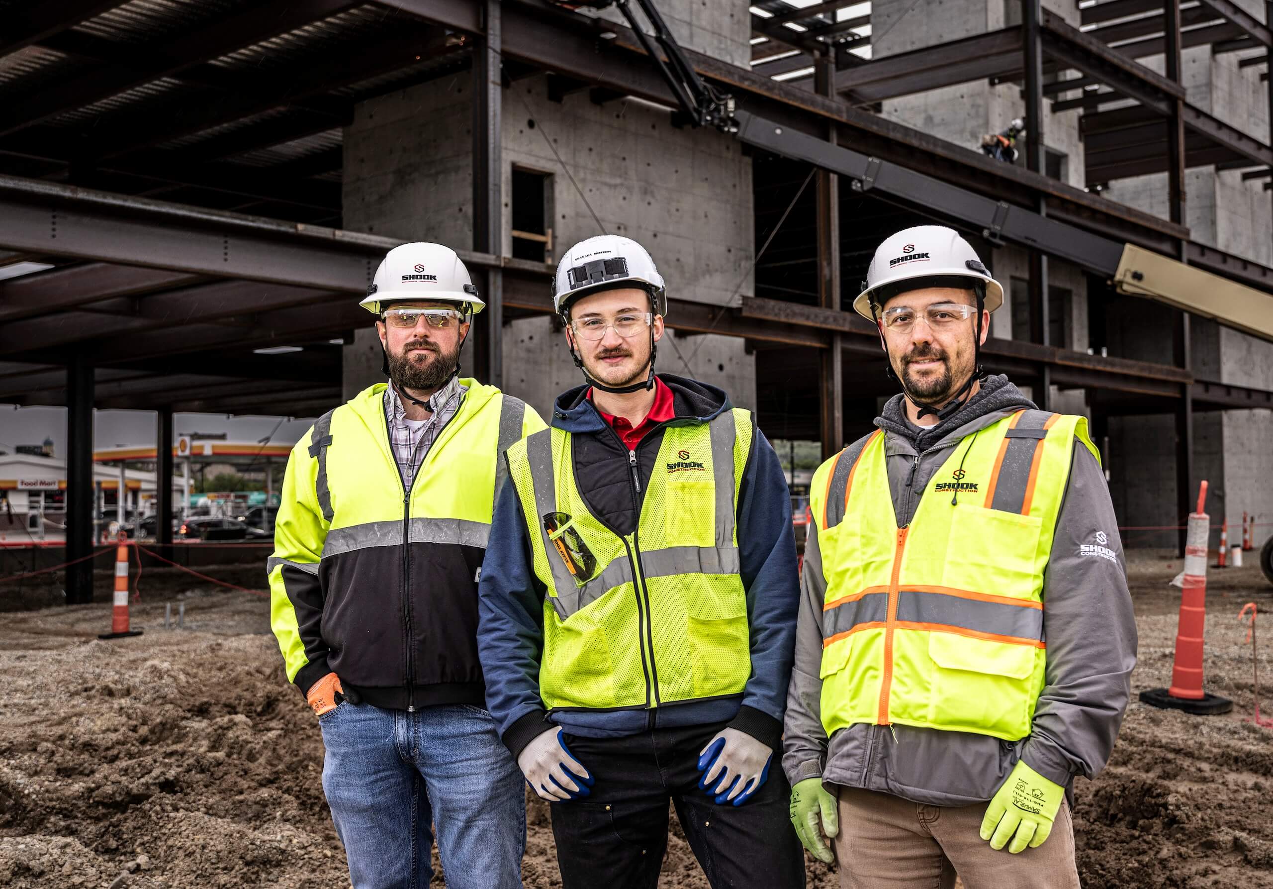 Three construction workers wearing safety vests, hard hats, and gloves stand at a building site with a steel-frame structure in the background.