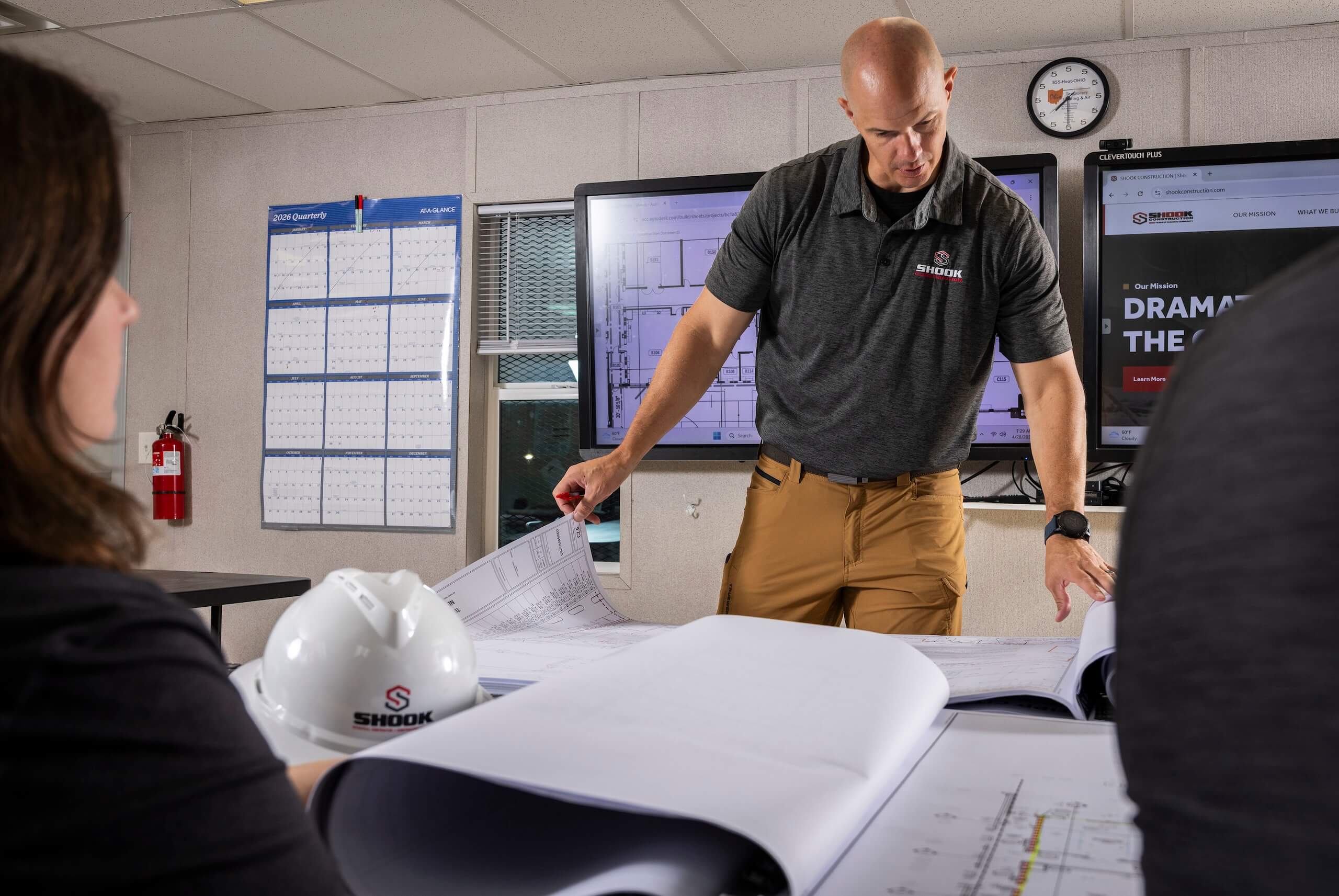 A man in a gray polo reviews blueprints with two colleagues in an office. Monitors display diagrams, and a white hard hat with a logo sits on the table. A wall clock and calendars are in the background.