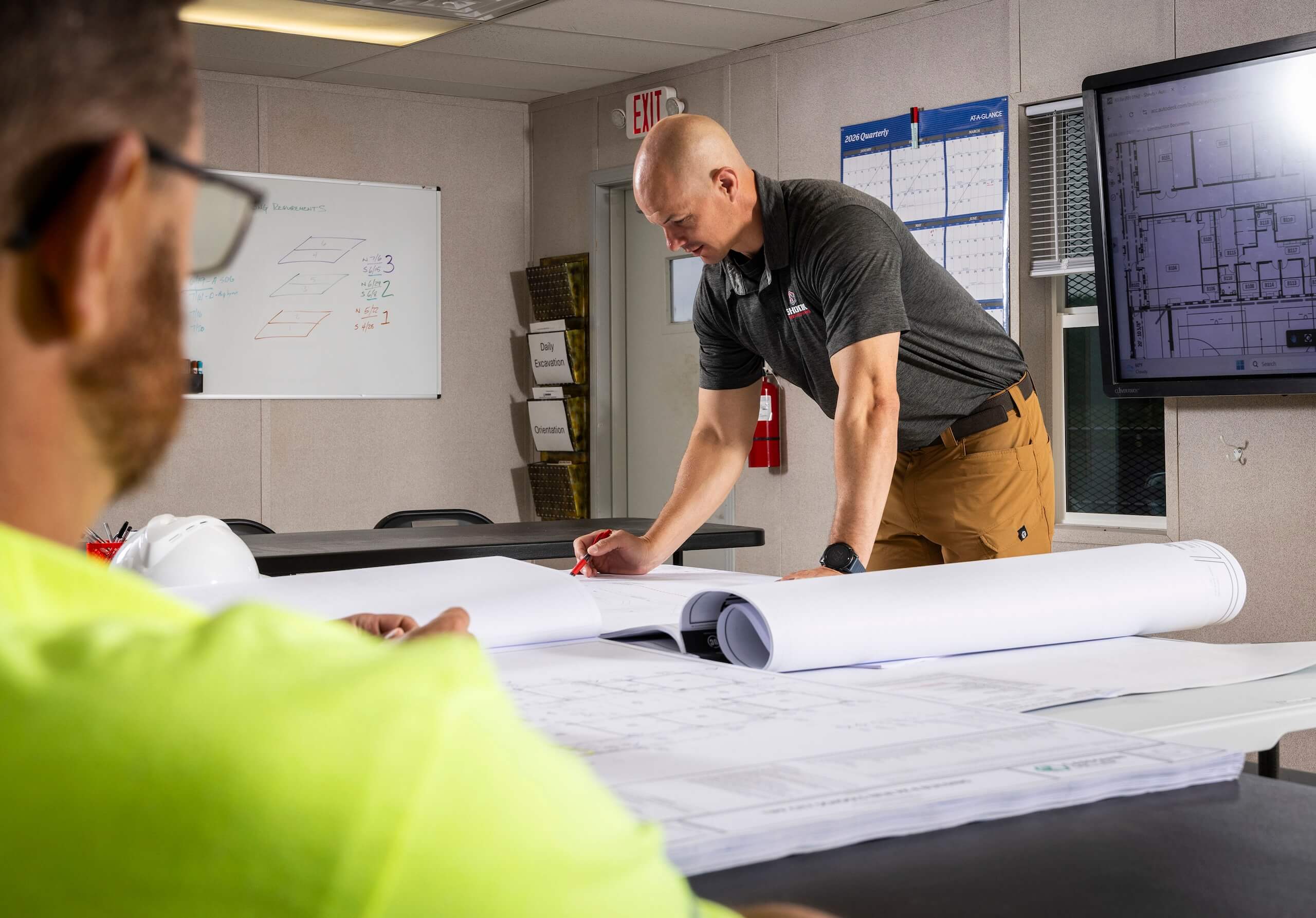 Two men review large blueprints in an office. One stands and writes on the plans while the other sits in the foreground. Walls display calendars, a whiteboard, and a screen showing building layouts.
