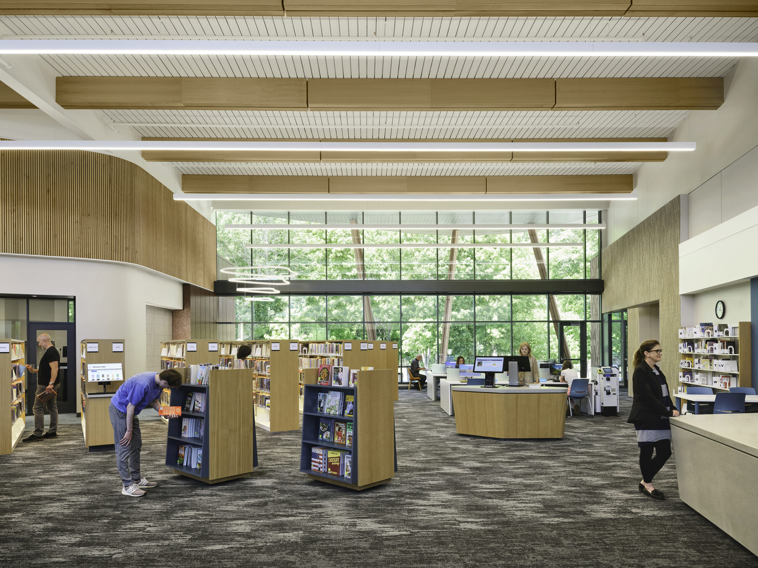 A modern library interior with large windows, bookshelves, and people browsing or working at desks. The space is well-lit with natural light and overhead fixtures, featuring wood accents and a carpeted floor.