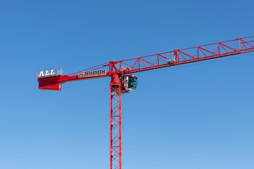 A red construction crane with ALL and SHOOK signs extends horizontally against a clear blue sky.